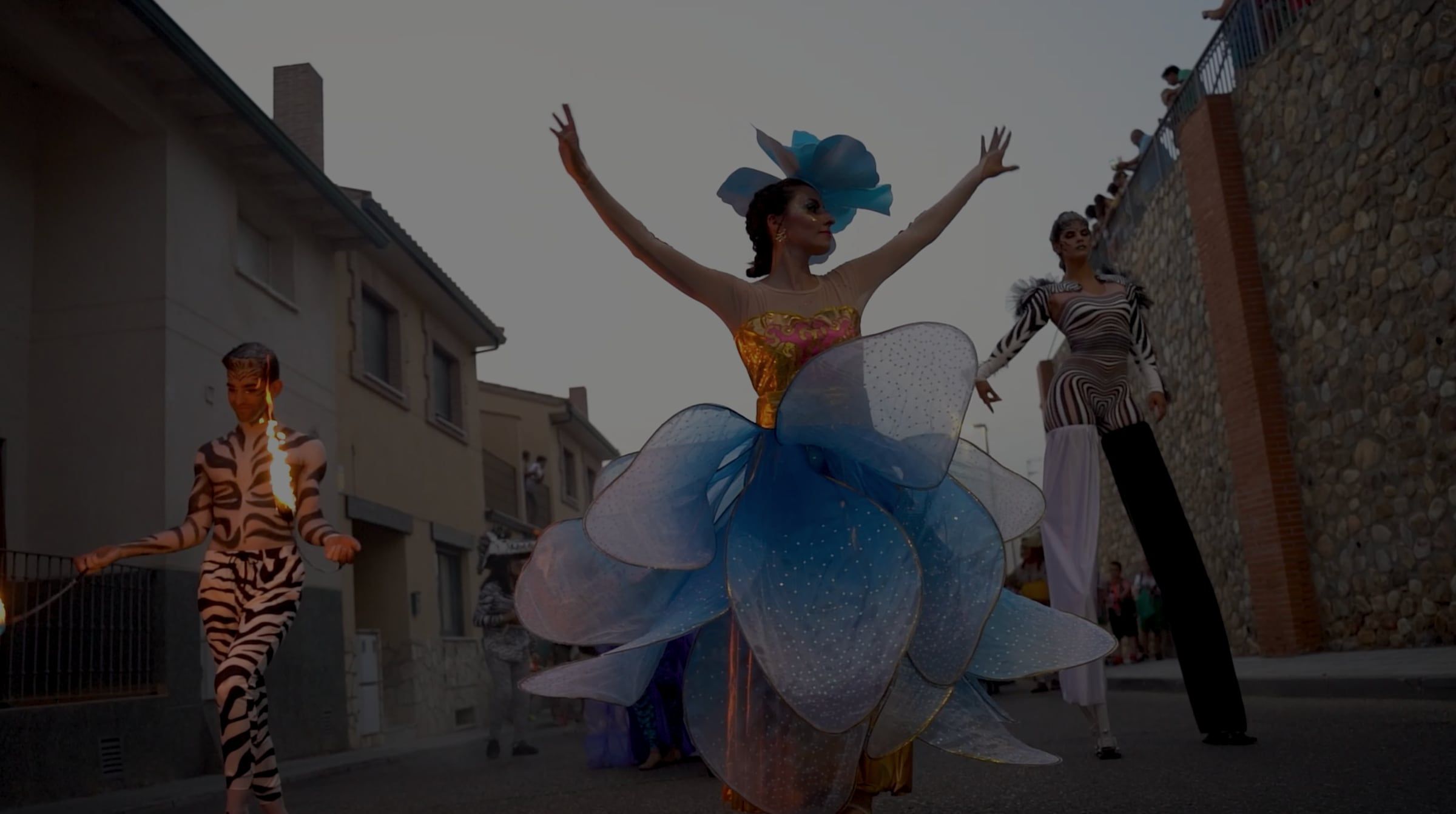 Pasacalles al atardecer con una bailarina vestida de flor azul, un malabarista de fuego y una zancuda con traje de cebra